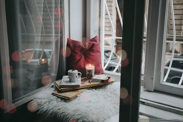 Idyllische Szene. Ein offenes Fenster. Ein rotes Kissen liegt darangelehnt. Darunter liegt eine flauschige weiße Decke. Auf der Decke steht ein kleines Tablett mit einer Tasse Kaffe und einer Kerze darauf