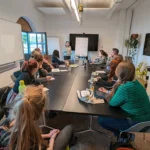 Viele Personen sitzen an einer Tafel mit Blick zu einem Flipchart und hören zu